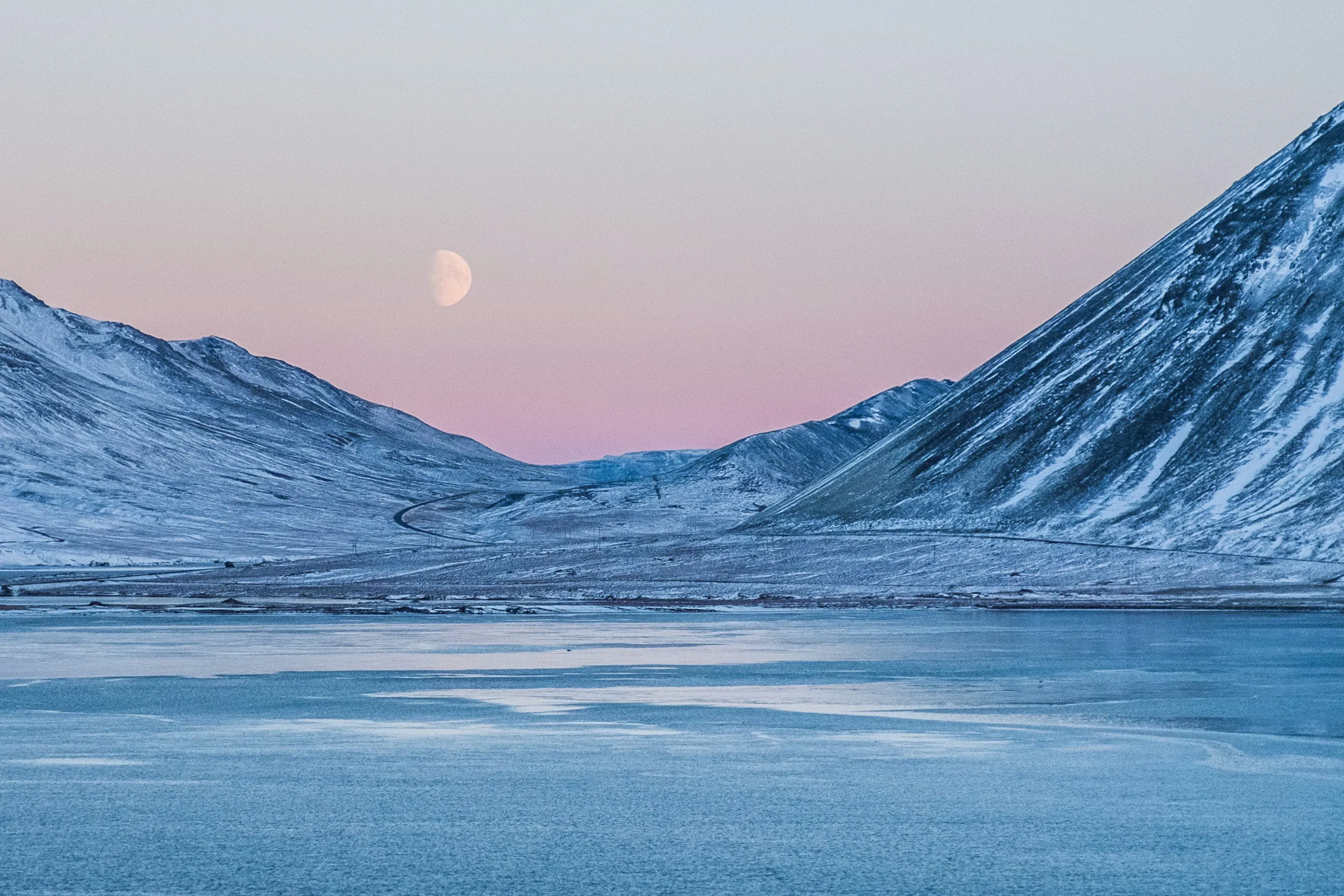 Snow-covered Arctic mountains under a pink twilight sky with moon rising over a frozen fjord